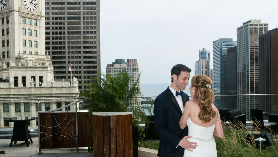 A newlywed couple standing on a rooftop terrace, posing against an urban skyline with historic buildings in the background.