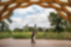 Couple kisses under a wooden archway in a park with city skyline in the background. Bright, sunny day with scattered clouds and greenery.
