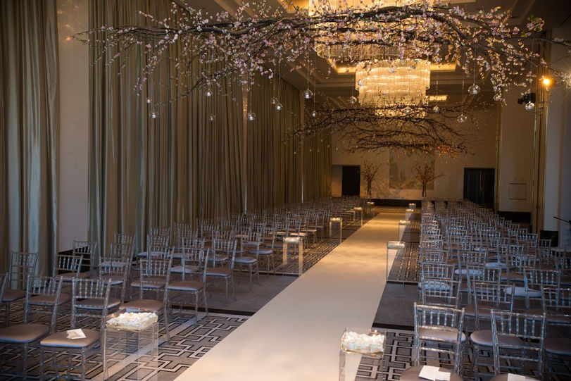 An elegant wedding aisle lined with clear chairs and decorated with floral arrangements hanging from the ceiling.