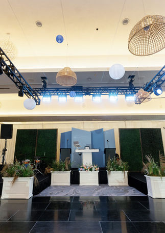 Wide view of the Radisson Blu Aqua Hotel Chicago ballroom stage decorated with a greenery wall backdrop, blue geometric panels, white planter boxes, blue and white hydrangea floral columns, and hanging woven rattan pendants overhead.