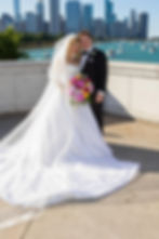 Bride and groom smiling by a waterfront with city skyline; bride in a white gown holding vibrant flowers under a clear blue sky.
