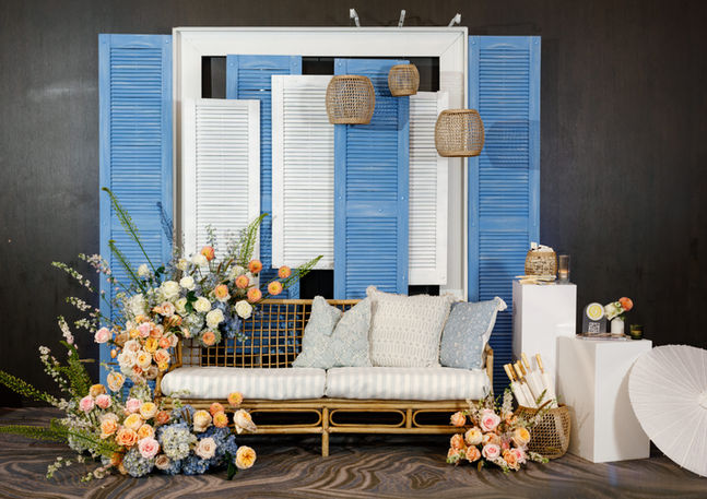 Full view of a styled lounge vignette at the Radisson Blu Aqua Hotel Chicago featuring a rattan loveseat with blue and white cushions, peach and blush floral ground installations, hanging woven pendants, and a blue and white shutter backdrop.
