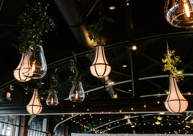 Close-up of Theater on the Lake Chicago's distinctive pendant lanterns and globe string lights hanging from the venue's green steel arched ceiling, interwoven with trailing greenery installations.