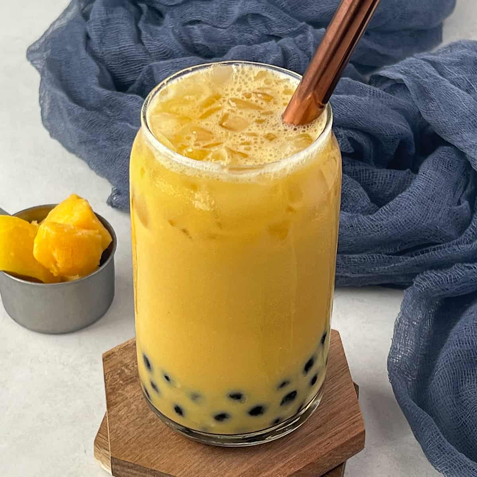 Yellow bubble tea with black tapioca pearls in a glass, metal straw, next to mango chunks in a cup. Blue cloth background.