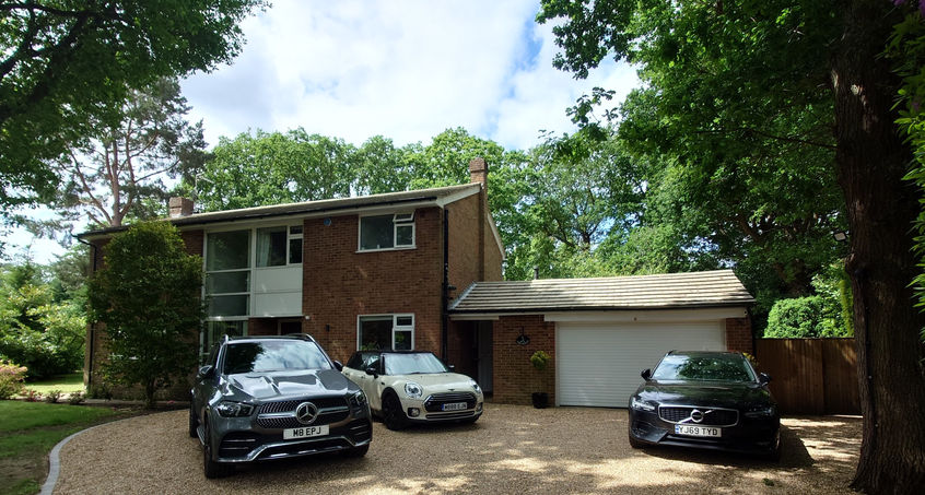 Front view of a two-story red-brick house with large windows and an attached white garage, a curved gravel driveway set among tall trees and greenery.