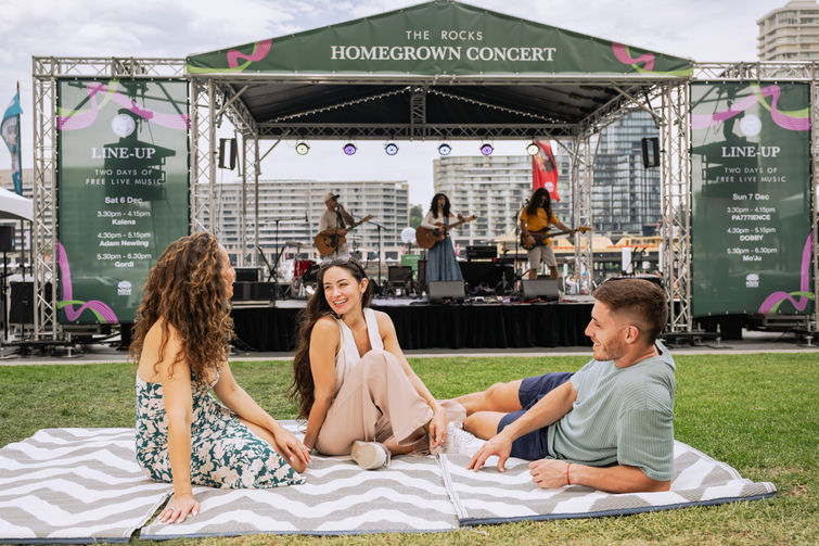 A Lifestyle photo of  2 women and a man on a picnic rug in front of a concert stage
in The Rocks, Sydney Australia
