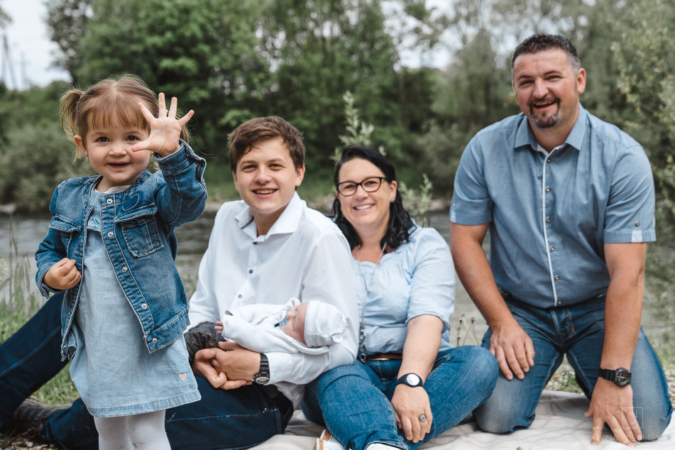A family of five enjoys the outdoors by a river. A young girl in a denim jacket waves at the camera, while her brother cradles a newborn. Their parents sit nearby, all beaming. Captured through NW Photography's lens, this moment reflects the art of professional portrait photography amidst natures greenery.