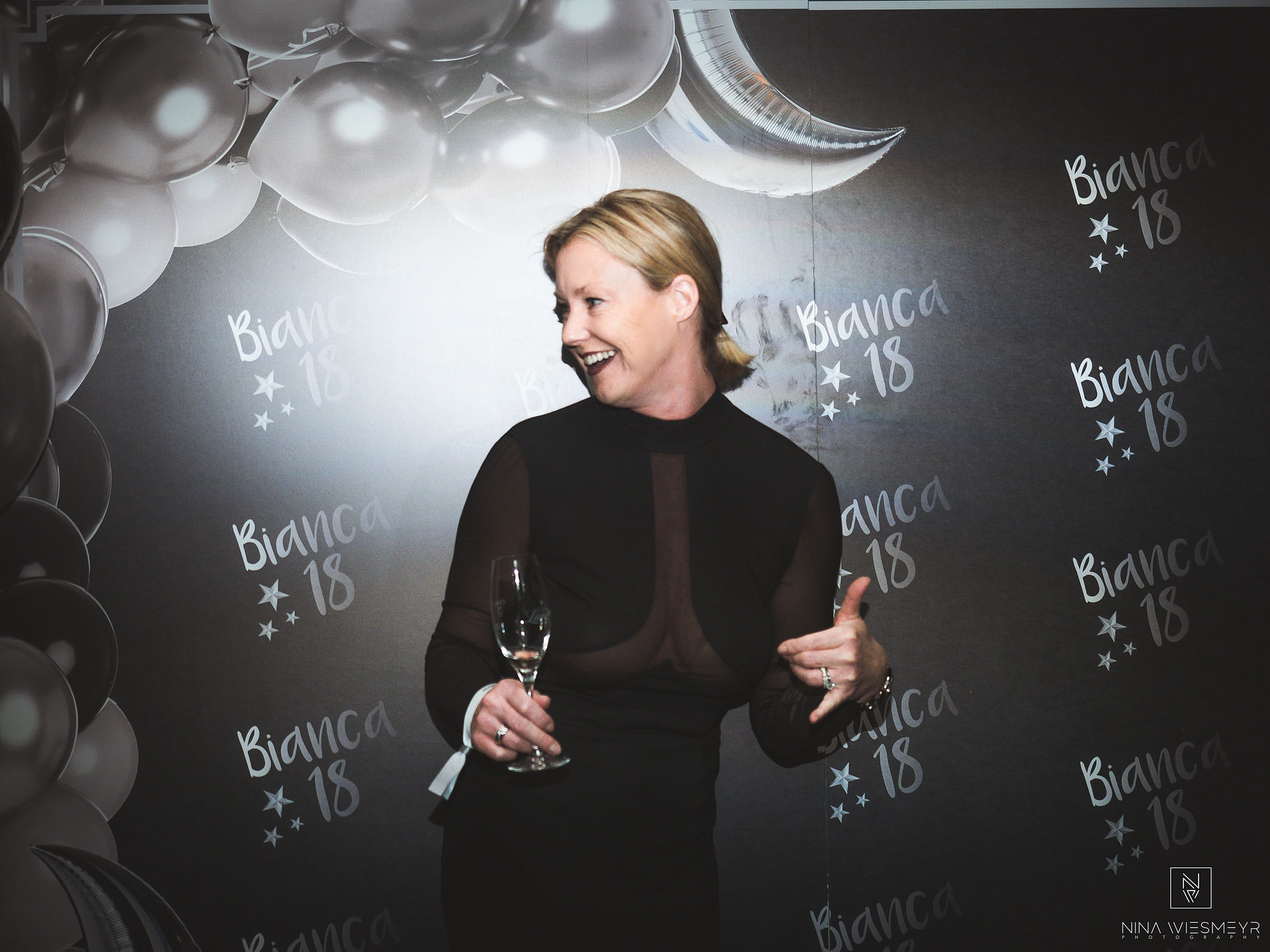 Eine Person in einem schwarzen Kleid lächelt mit einem Glas Wein auf einer Veranstaltung, festgehalten mit hochwertiger Eventfotografie. Im Hintergrund ist ein Banner mit der Aufschrift „Bianca 18“ zu sehen, ergänzt durch festliche Ballondekorationen.
