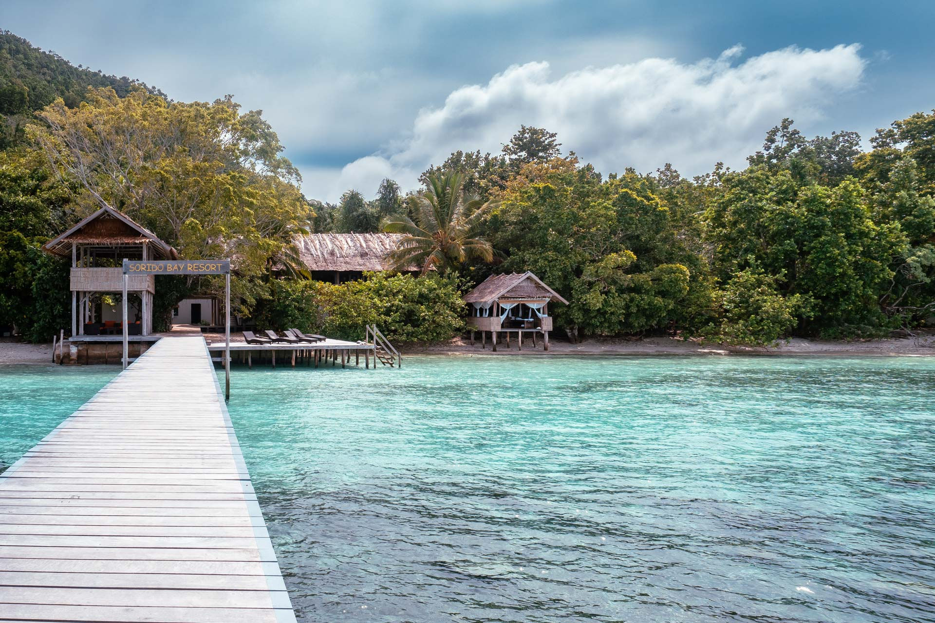 A wooden pier extends over the crystal-clear turquoise waters of Raja Ampat, leading to a tropical island with lush greenery and rustic beach huts nestled among the trees under a partly cloudy sky—a true Divers Paradise.