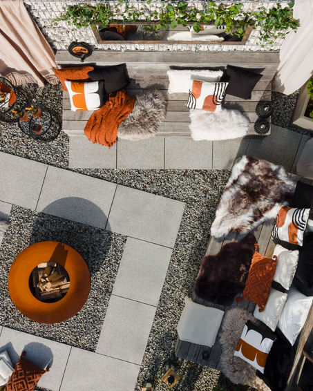 Aerial view by a professional commercial photographer showcases a stylish outdoor patio with cushioned seating, patterned pillows, and cozy throws. A circular fire pit rests on paved gravel, surrounded by lush greenery, creating a warm and inviting atmosphere.