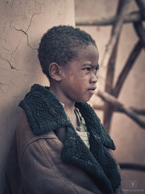 A young boy stands against a wall, looking pensively to the side. Captured by professional people photographer Nina Wiesmeyr, he wears a large, heavy coat and has short, curly hair. The rustic backdrop adds depth to this evocative piece of portrait photography.