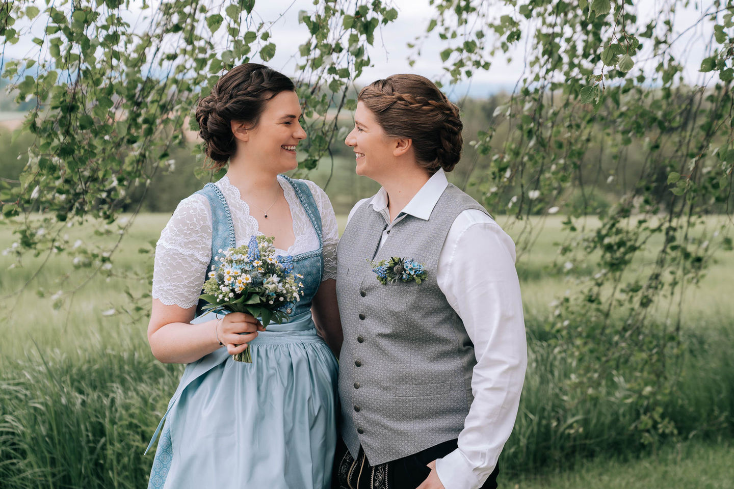 Amidst a grassy sanctuary with trees and a cloudy sky, two people stand smiling at each other. One wears a traditional dress holding a floral bouquet, while the other sports a vest and white shirt—an exquisite example of NW Photography capturing real emotions in stunning portraiture.
