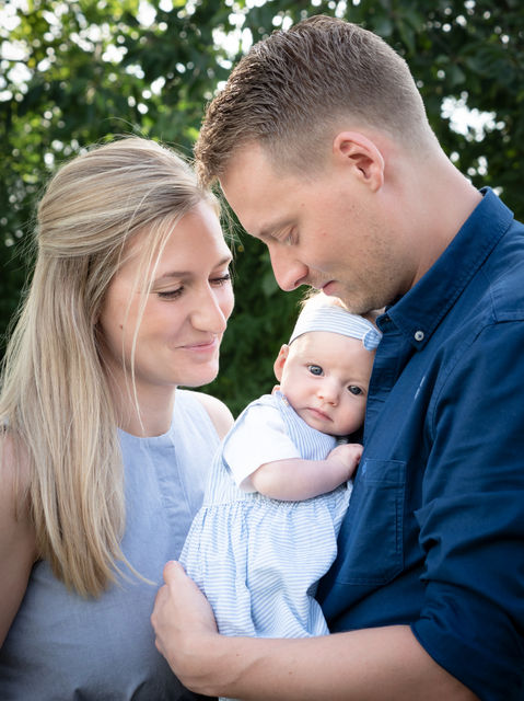 A smiling couple embraces a baby wrapped in a blue-striped outfit and matching headband. The woman has long blonde hair, and the man has short light brown hair. Captured by an NW Photography professional portrait photographer, they are outdoors with lush green foliage in the background.