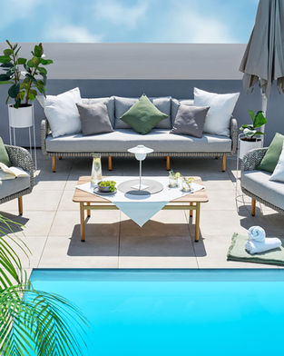 A modern outdoor patio with wicker furniture, including gray cushioned sofas and chairs around a wooden coffee table. Captured by a professional commercial photographer, the table holds a lamp, drinks, and snacks. Fresh towels and a closed umbrella are near a bright blue pool under a clear sky.