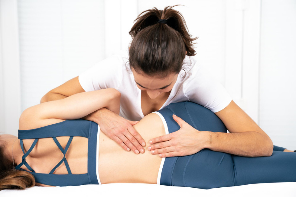 A physical therapist, captured by professional business photographer Nina Wiesmeyr, assists a person in blue athletic wear who is lying on their side. The therapist, wearing a white shirt, applies pressure to the persons lower back. The softly lit background emphasizes the supportive interaction.