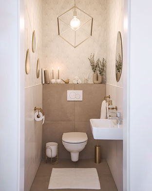 A modern bathroom perfect for product photography, featuring a wall-hung toilet, geometric pendant light, and ambient candles. Beige tiles and gold fixtures enhance the white sink. Round mirrors and greenery complete the decor, with a white mat on the floor adding a professional touch.