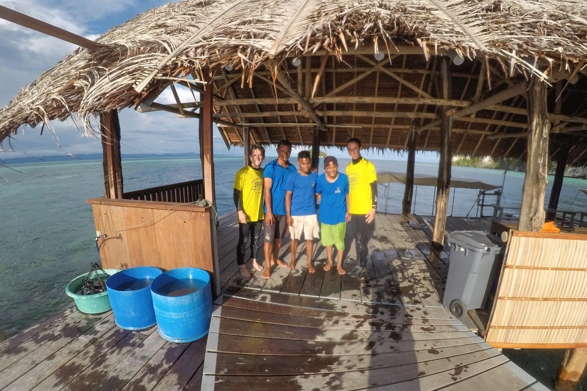 Under a thatched-roof structure in Raja Ampat, five people stand casually on a wooden deck beside the crystal-clear water. With two blue barrels and a gray bin nearby, this divers paradise offers endless exploration under partly cloudy skies. Perfect for any travel blog entry.