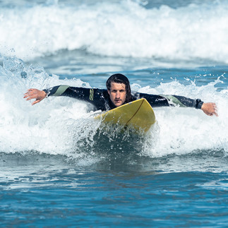 A surfer in a black wetsuit skillfully paddles on a yellow surfboard through white waves in the blue ocean, captured expertly by NW Photography. His arms extend outward as waves splash around him, creating a dynamic and captivating scene without the sky in view.
