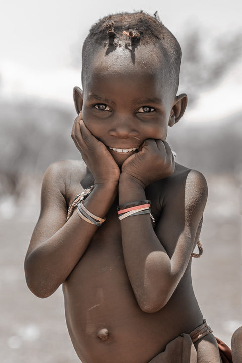 A child smiles warmly, chin resting in both hands. Adorned with multiple bracelets and a distinctive hairstyle, their joyful expression is the highlight of this soft-focused scene. This image is a perfect example of NW Photography by Nina Wiesmeyr, capturing real emotions through stunning people photography.