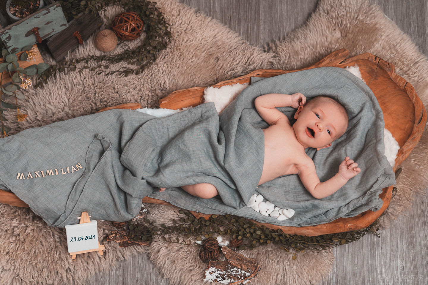 A newborn baby lies swaddled in a gray blanket on a rustic wooden bed with plush fur underneath. The name Maximilian is spelled out with wooden letters, and a small easel displays the date 29.04.2024. This setup beautifully showcases the art of professional portrait photography, surrounded by natural decor.