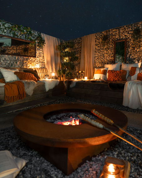 Cozy outdoor seating area at night, captured by NW Photography, features a central fire pit. Surrounding benches are adorned with pillows and blankets in earthy tones. Soft ambient lighting from candles and lanterns enhances the warm, inviting atmosphere under a starry sky.