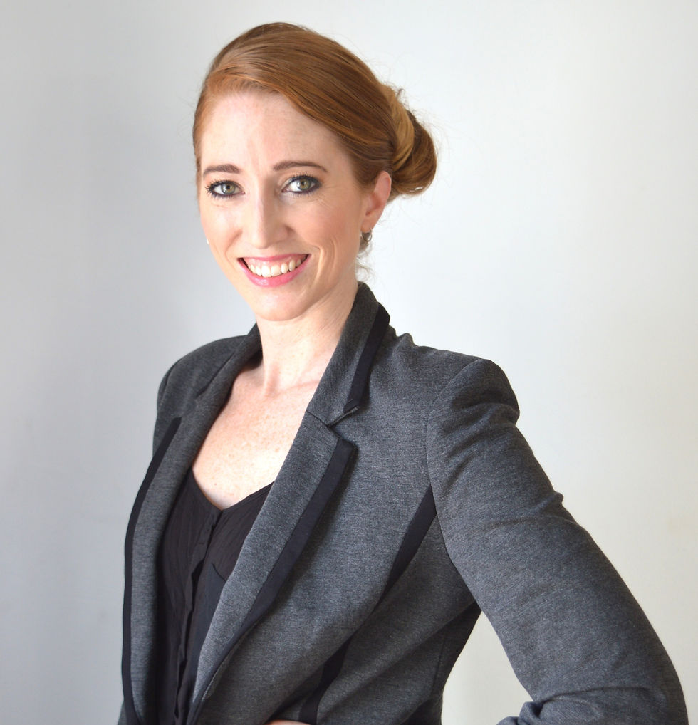 Smiling woman with red hair in a bun, wearing a gray blazer over a black top against a plain light background. Confident and professional mood.