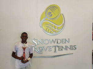 Young athlete, dressed in white, holding a trophy stands smiling by a sign reading "Snowden Grove Tennis" with a yellow logo on a white wall.