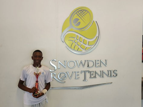 Young athlete, dressed in white, holding a trophy stands smiling by a sign reading "Snowden Grove Tennis" with a yellow logo on a white wall.