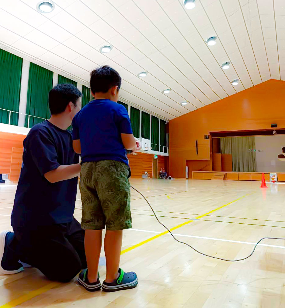 室内体育館で男の子がリモコンを持ち、膝をついている男性と一緒に車を操縦している。明るい白い天井と緑のカーテン。ドローンクロスがジュニアドローン教室を開催。