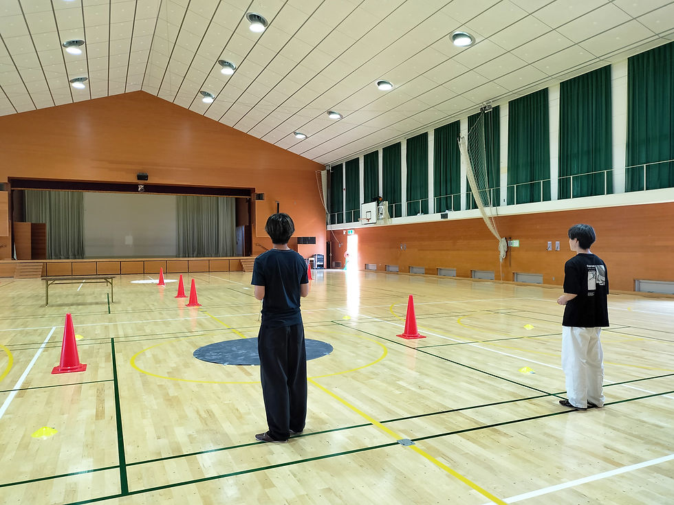 体育館で2人が向かい合い、床に並べられた赤いパイロンの間に立っている。木製の床と緑のカーテンが背景。静かな雰囲気。ドローンクロスがジュニアドローン教室を開催。