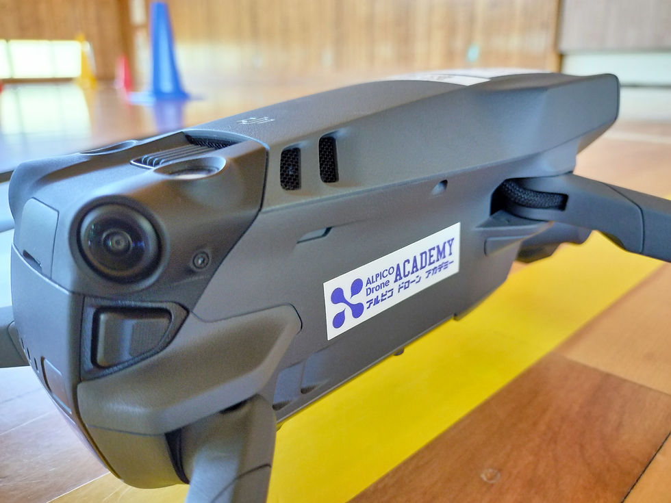ドローンが木製の床に置かれ、ボディに「ALPICO Drone ACADEMY アルピコ ドローン アカデミー」のラベルが見える。背景にはカラフルなコーンがある。ドローンクロスが定点撮影と高高度撮影を実施。
