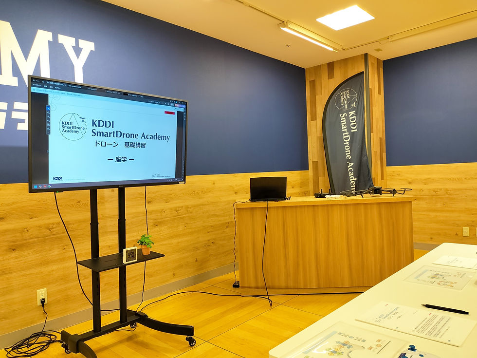KDDI SmartDrone Academyの教室。スクリーンに「ドローン基礎講習」と表示。木目調の壁に机とノートPC、資料が並ぶ。ドローンクロスがジュニアドローン教室を開催。