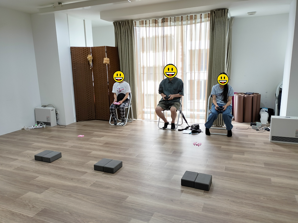 3人が椅子に座り、リモコンを持ち笑顔。背景はカーテンと木製パネル。床にクッションブロックが並ぶ。リラックスした雰囲気。ドローンクロスがジュニアドローン教室を開催。