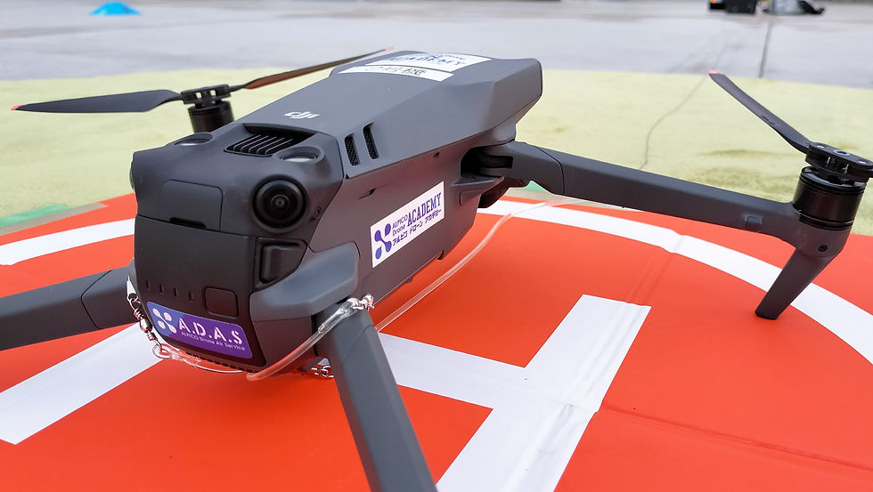 オレンジ色のHパッド上にある黒いドローン。側面に「ADAS」と「ACADEMY」のステッカーが貼られている。背景は空港のような屋外。ドローンクロスが定点撮影と高高度撮影を実施。