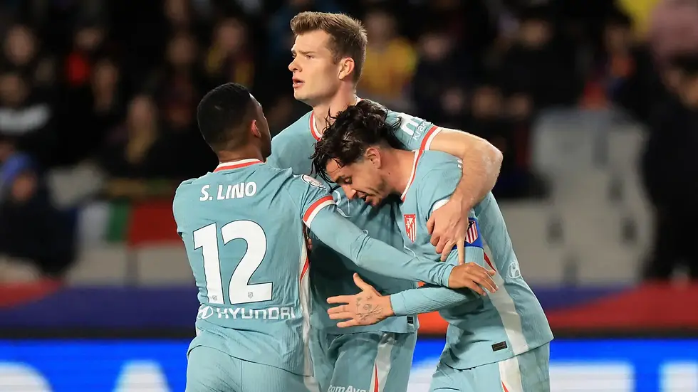 Three soccer players in light blue jerseys embrace on the field, celebrating. The background shows a blurred crowd. The mood is joyful
