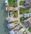 Drone top-down view of a waterfront neighborhood showcasing homes with private docks, pools, and green lawns along a calm shoreline. Captured by Strata Visuals.