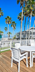 Bright outdoor deck area featuring white patio furniture, wooden flooring, and palm trees under a clear blue sky — perfect tropical retreat captured by Strata Visuals for Vacation Rental Photography.