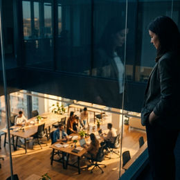 A founder stands alone at a glass wall on an upper floor, looking down at a team working two levels below. The image carries the quiet distance growth creates between a leader and the room, the moment a leader can still see the team but is no longer in the conversation.