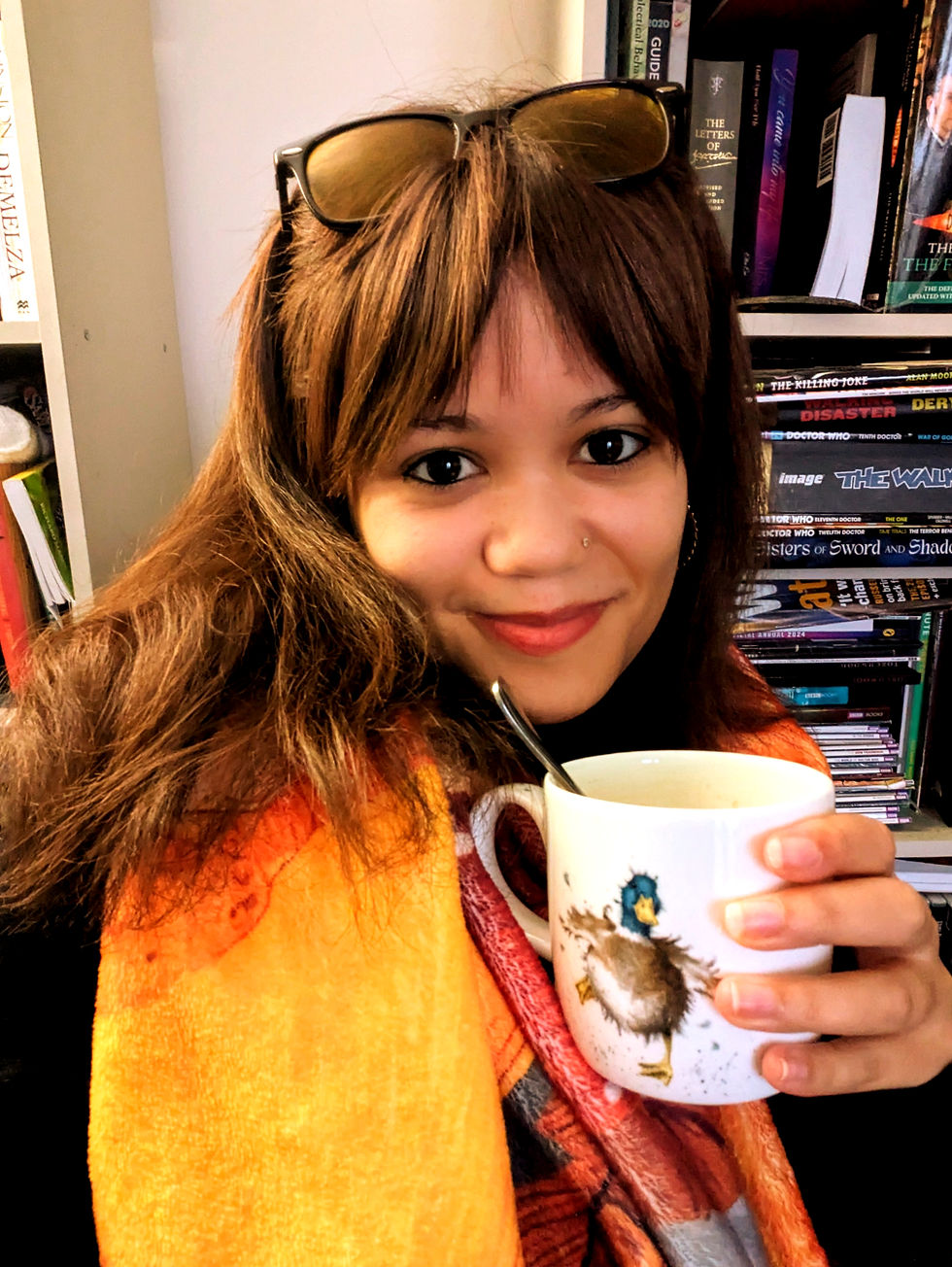 A woman smiling at the camera holding a duck cup with a bookshelf behind her. This woman works as a book editor in the UK and offers affordable book editing services.