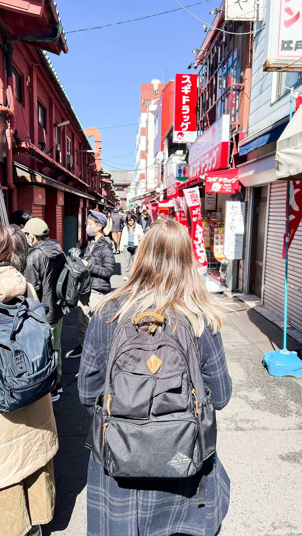 a woman with long blonde hair walks down an alley with small shops in Japan, her back is to the viewer. She's wearing a backpack and coat.