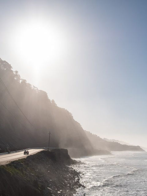 The sun shining over a ridge leading down into the shore. In the distance, a car drives down a road.