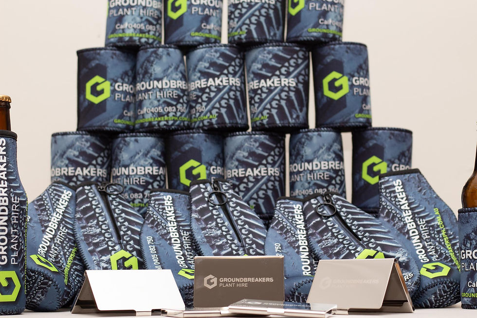 Stacked blue koozies with Groundbreakers logo, business cards, pens, and brown bottle. Logos in green and white text. Indoor setting.