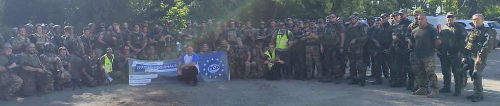 Un gruppo di giocatori di softair, vestiti con uniformi mimetiche ed equipaggiamento tattico, si riunisce in un'area boschiva. Alcuni portano fucili ad aria compressa (ASG) e radio. L'immagine cattura un momento di aggregazione e preparazione durante un raduno o un evento sportivo.