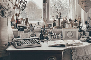 Cozy art studio with typewriter, open sketchbook, dried flowers, brushes, and jars on a wooden desk by a window. Neutral tones create a calm vibe.