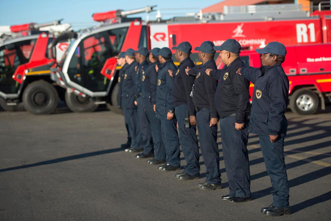 Firefighters standing in formation with red fire trucks at or tambo airport