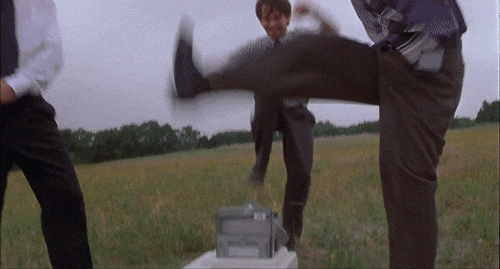 Three people in suits aggressively smash a printer in a field, expressing frustration. The background shows grass and trees.