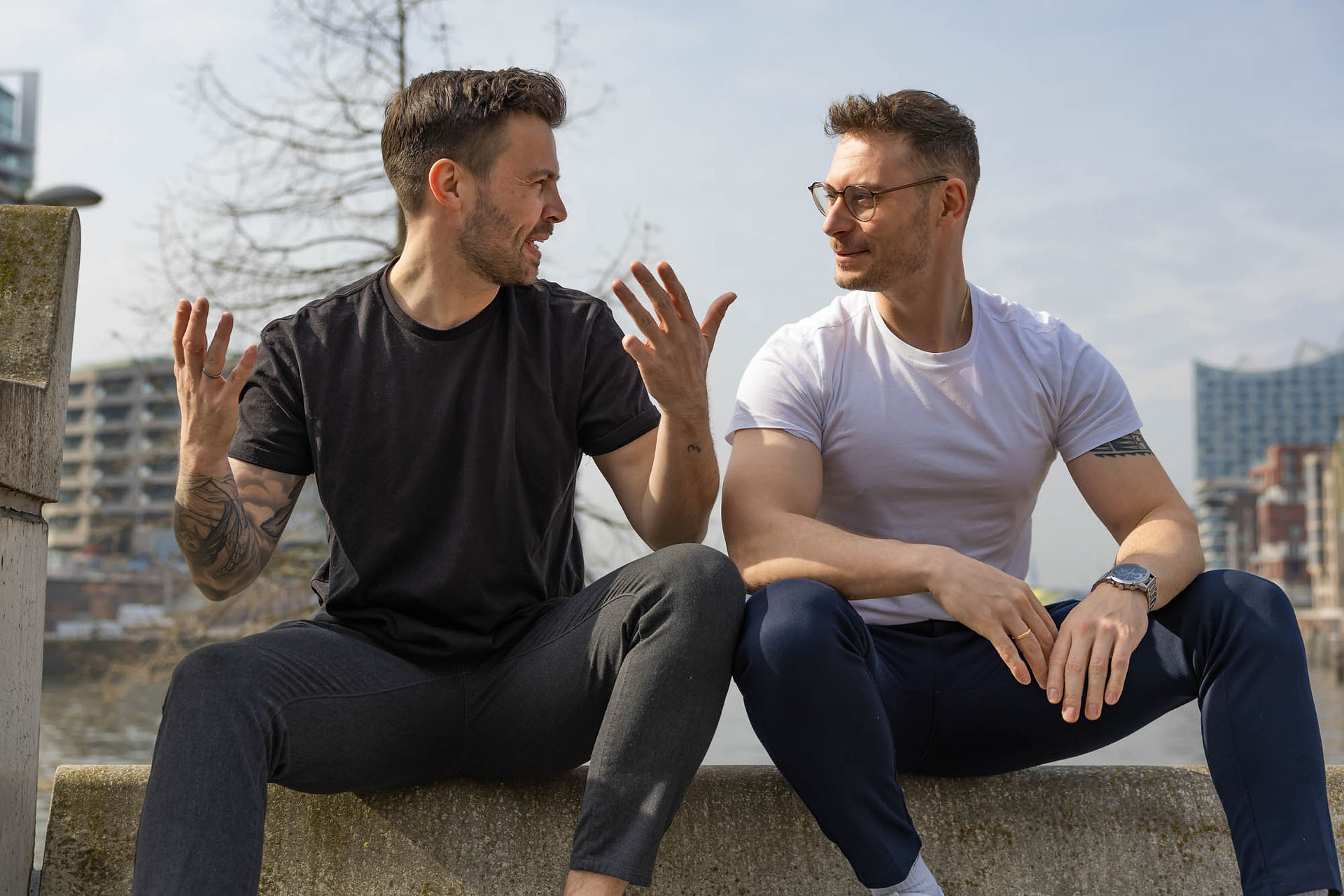 Zwei Männer im weißen und schwarzen Tshirt sitzen auf einer Mauer am Wasser in der Hafencity.