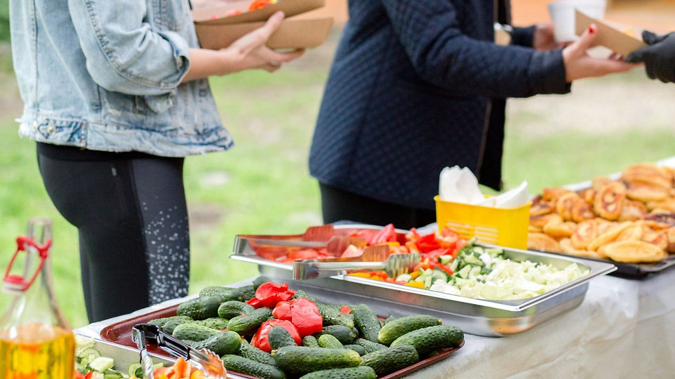 Outdoor buffet with people serving food. Trays of cucumbers, tomatoes, pastries on a table. Green grass in the background. Casual setting.