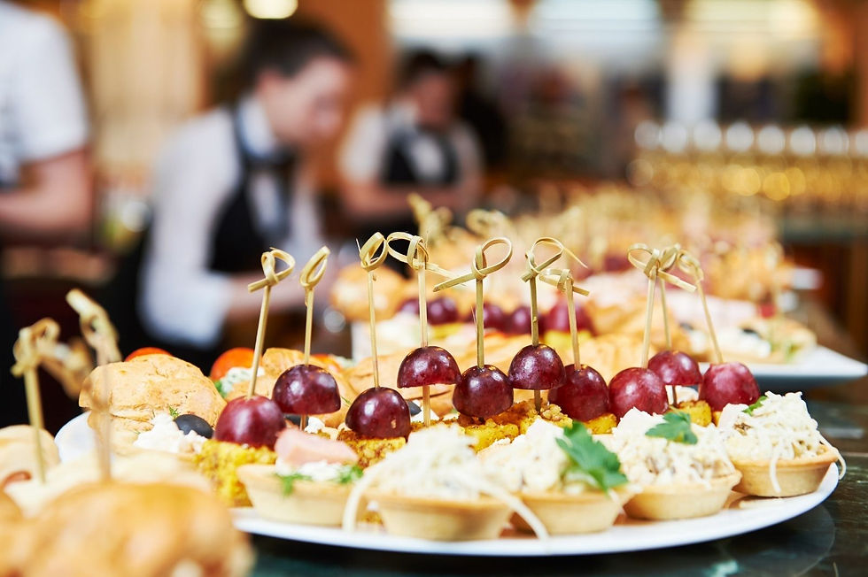 Buffet trays of bite-sized appetizers topped with grapes, with blurred chefs in a warm restaurant kitchen behind.