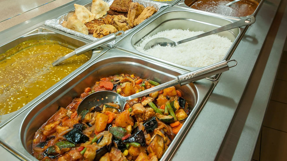 Buffet trays of white rice, curry, vegetable stir-fry, and fried snacks with serving spoons in a stainless-steel cafeteria.